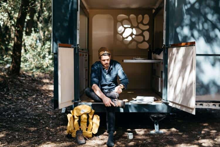 Man sitting at the doorstep of a portable tiny house, tying his shoelace with a backpack on the floor next to him and a forest peeking at the background