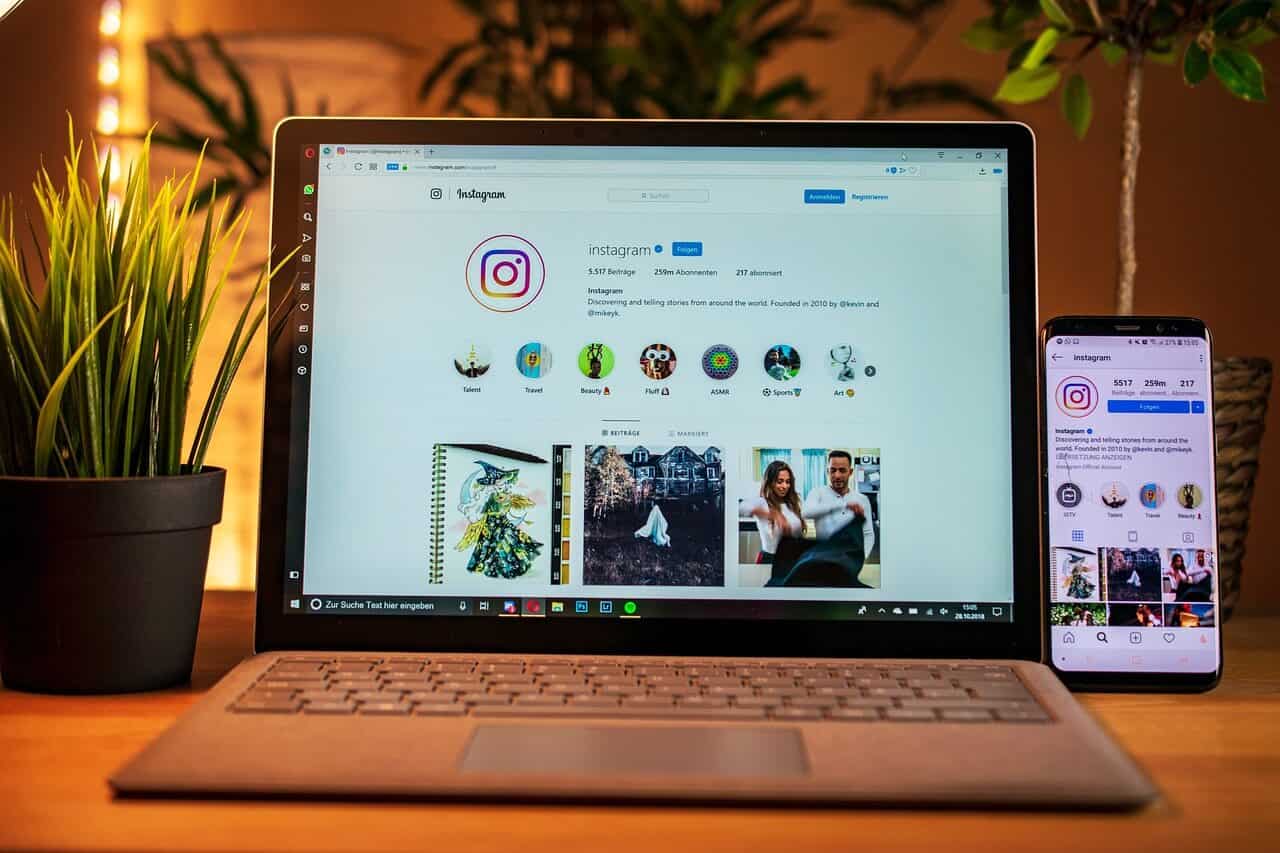 A laptop and phone screen side by side showing Instagram's official Instagram account. On the left side, there's a grassy pot plant. The back is also filled with leafy pot plants.