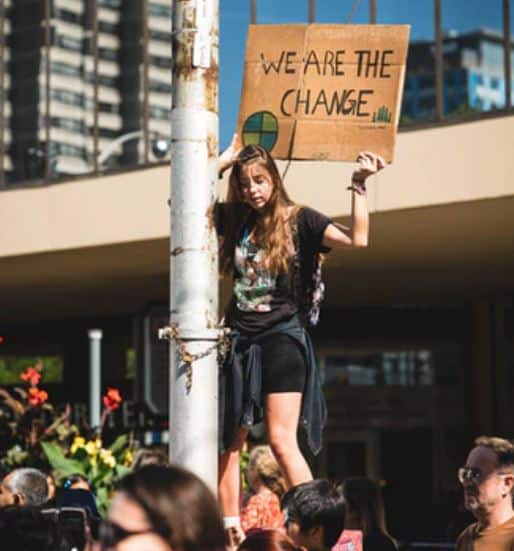 Young girl climbing a street light post and looking down. She's at some sort of protest. She's holding up a cardboard sign saying "we are the change" with a globe-like ball green and blue 4-piece pie chart in the left corner.
