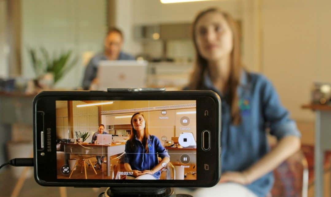 Blurry background and a phone screen at the forefront. On the screen we see that it's a woman recording herself on video in an office, with her coworker sitting at the desk with her laptop on the back right of the woman whose being recorded.