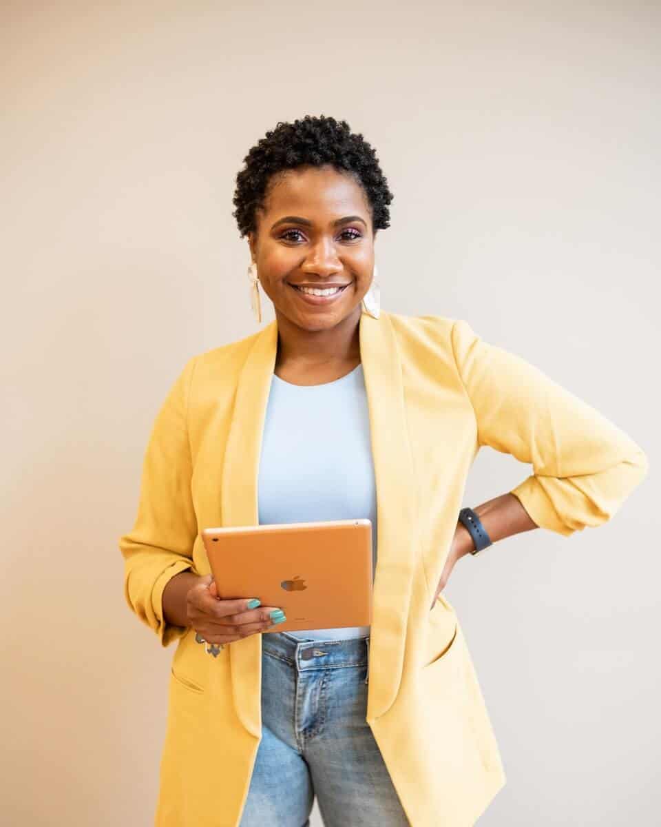 Woman in yellow blazer holding yellow ipad, with short black curly hair and her left hand at her hip. She's looking at the camera and smiling.