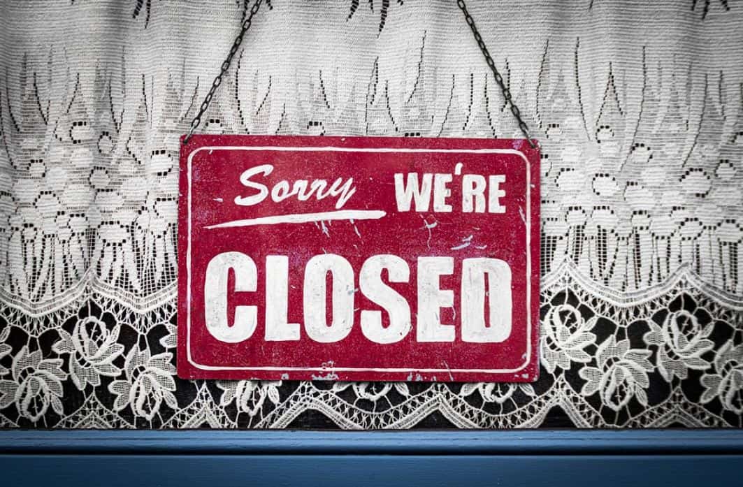Red sign hanging on a window with a blue pane and white mesh curtains, saying "Sorry, we're CLOSED".