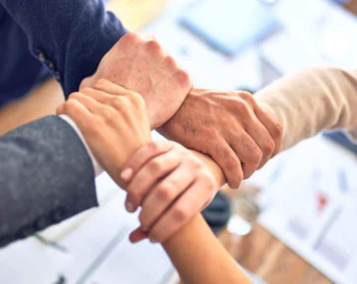 Four people cross-holding hands, aka holding each other's wrist in a chain so all 4 hands connect, in dress shirt and tuxedo sleeves, with paperwork on the table in the background, suggesting an office setting. Image chosen to represent building trust with borrowers.