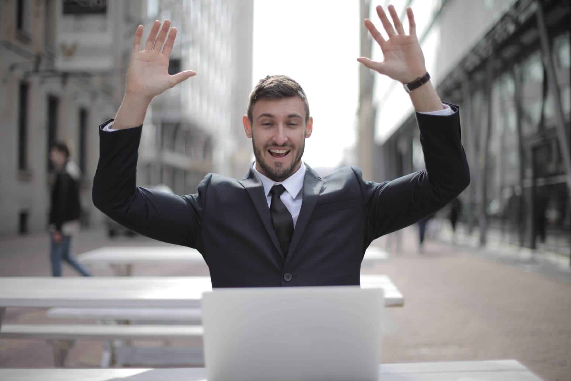 Man in suit sitting at a caffee outdoors with his laptop in front of him, smiling and cheering on with both hands raised in the air while he's looking at the screen.