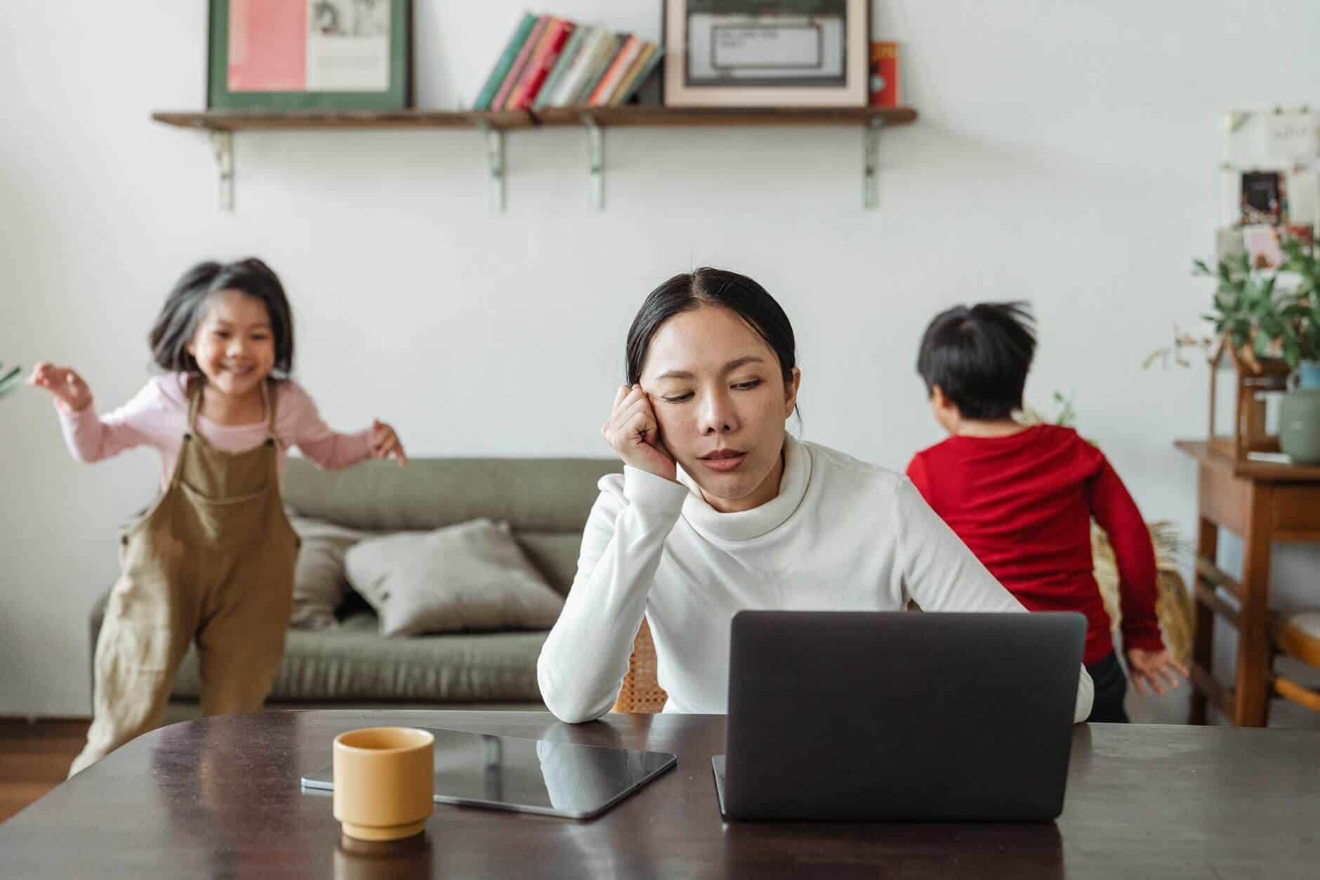 Woman sitting at her living room table in front of her laptop, with a coffee cup and her head rested on her right hand, looking tired and having a hard time focusing, and her two children happily running around in the background.