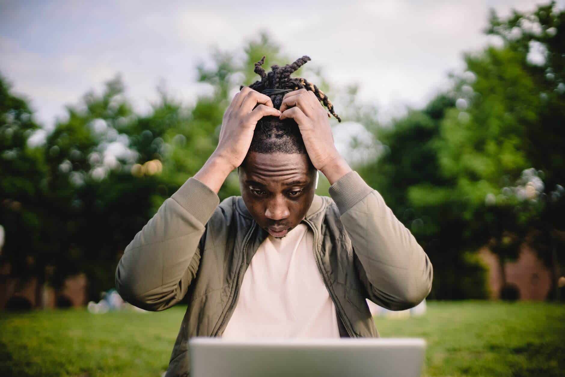 Man in a jacket with braids holding his hands on his head, with a worried and stressed look, gazing down at his laptop screen. He's working from a park bench.