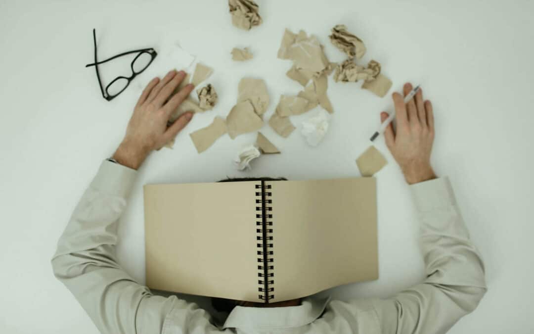 Person lying face flat on their desk, with an open notebook covering their head and paper shreds scattered on the desk. Their black glasses are in the upper-left corner of the image. Used to represent loan officer burnout.