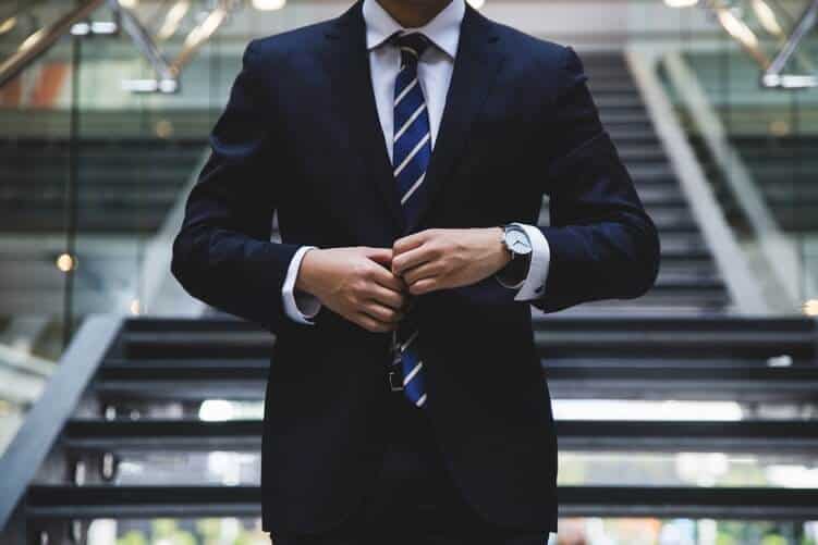 Main in suit adjusting his jacket, his watch on his left hand and a starircase behind him. We can only see his torso.
