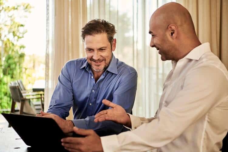 Man in blue and man in white button-up shirts sitting and looking at the laptop screen, smiling. The man in the white shirt is pointing at the screen with his hand. Seemingly office setting, with a small door leading to a patio on the left in the background.