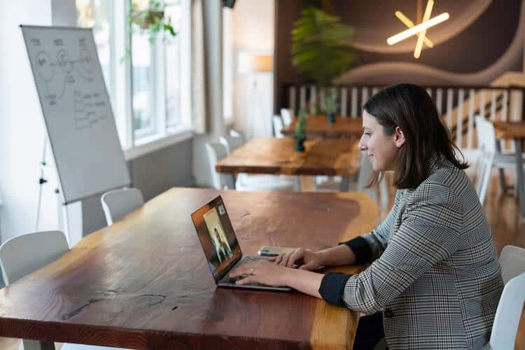 Womman sitting a a desk in what looks like a coworking space - decorations and more desks in the back, and a white paper board on the left next to the windows. She's on her laptop on a meeting call with someone.