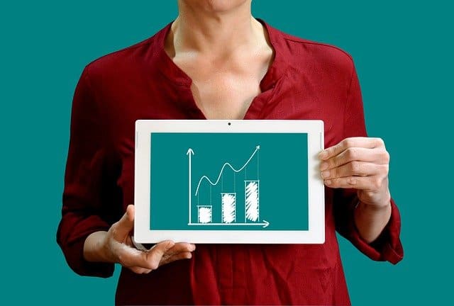 Person in red shirt visible from neck down on a cyan-green background showing a tablet with a white border and matching cyan-green background on the image on the screen, showing a sales chart, with 3 stacks higher to the right and a line with an arrow above going upwards, a mockup for a chart showing profit.