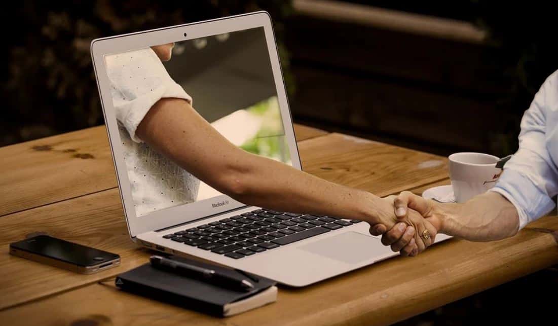 Edited image of hand reaching out from the laptop screen, shaking hands with the person sitting in front of the laptop. There is a notebook and a phone in addition to the laptop on the table, too.