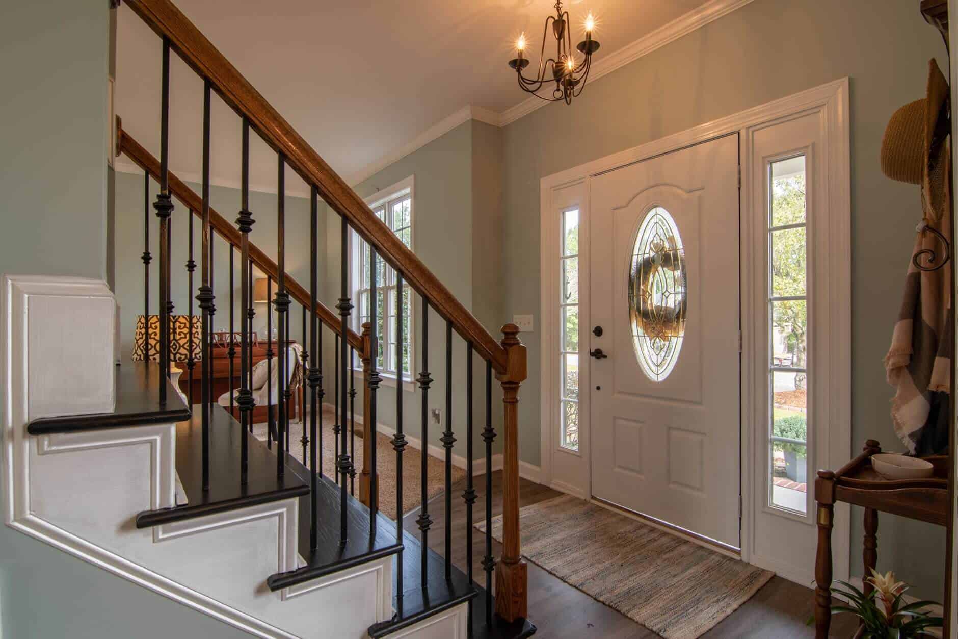 House entrence from the inside, with a brown laminate and brown stair case, some other wooden furniture and lamps peeking at the back. Next to the door is a small table and coat hange and a plant. Above the white door, there's a vintage chandelier. The walls are olive.