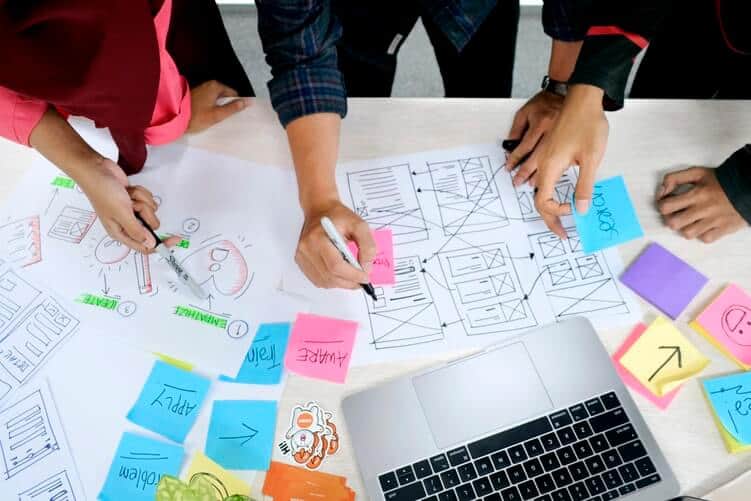 Design team sketching service icons and other assets in front of a laptop on the table. The image is from a birds-eye vew and we can only see the table, papers, pens, keyboards and hands, but not the screen or the heads of the people