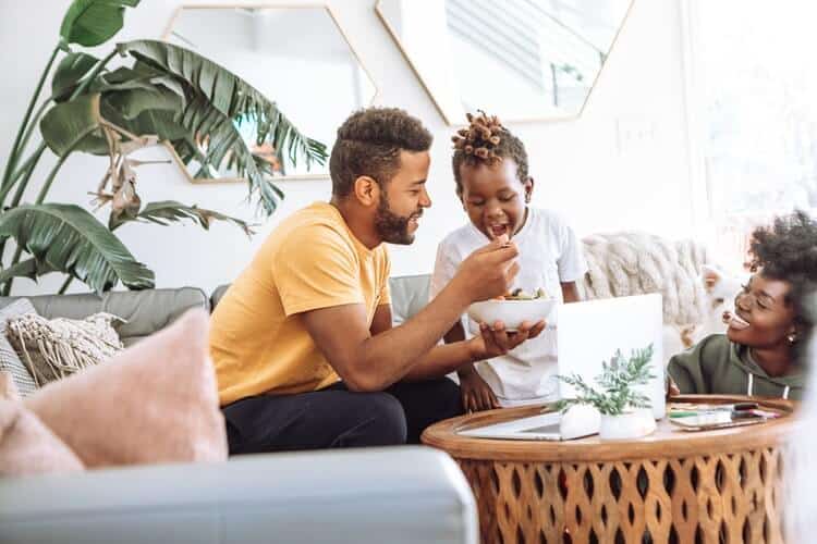 Family sitting at a woven living room table on a grey couch with pink pillows. The mother is crouching at the base of the table and laughing at the sight of the father feeding a food bowl to his laughing son. At the table, there is an open laptop, and a window in the back. Picture to represent happy home refinance opportunities.