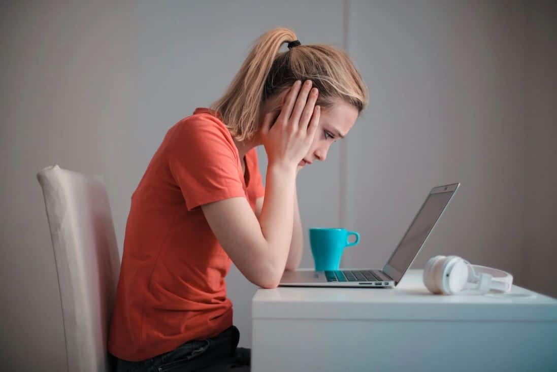 Worried Woman resting her head in her palms in front of her laptop in her white-furnished home office, with a blue coffee cup on the table next to her.