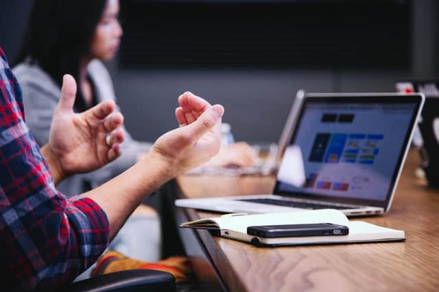 Person sitting at a table with other people, hand gesturing something. On the table in front of him, there's a laptop, notebook and a phone.